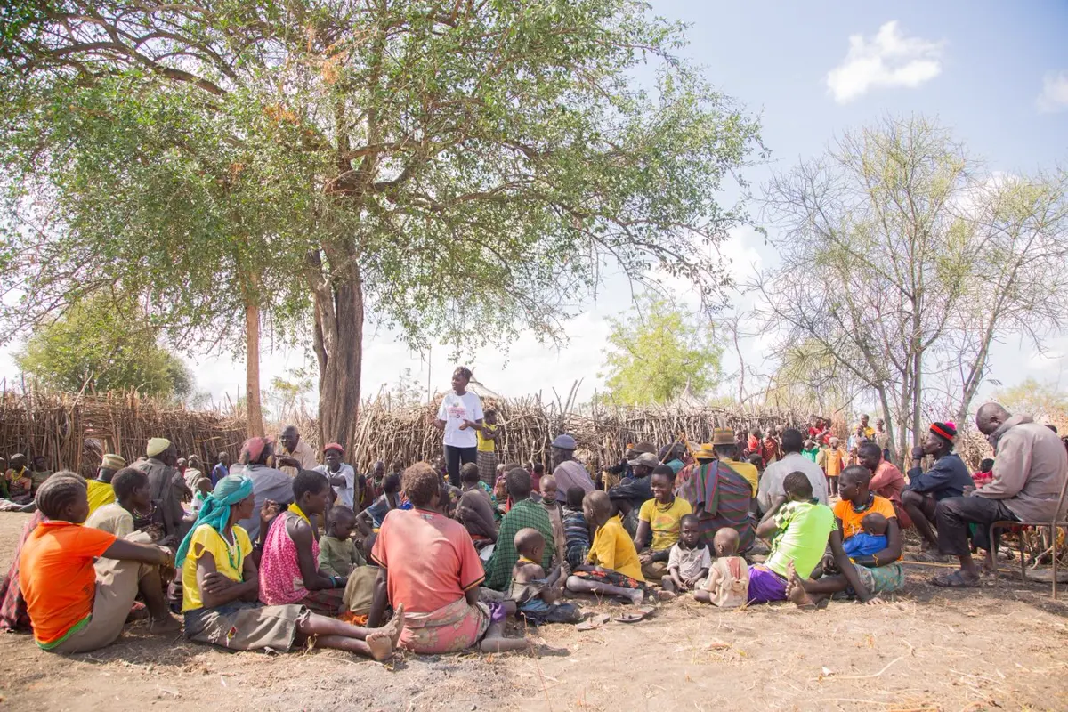 A large group of people seated together on the ground beneath a wide tree.