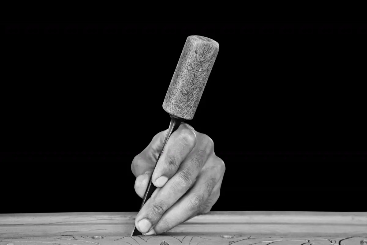 A hand holding a chisel against wood, lit against black.